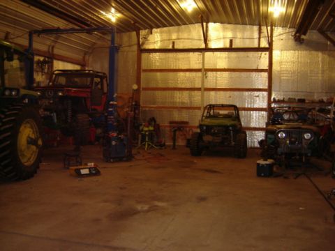 1992 Ford Bronco - Dimly lit metal pole-barn shop interior with a green farm tractor at left, a red-and-black truck body up on jack stands, a green side-by-si…