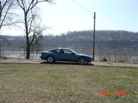 1993 Ford Probe - A teal coupe is parked on a dirt road next to a body of water under a clear sky.