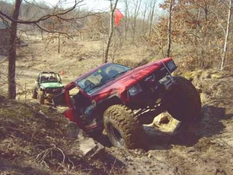 1992 Ford Bronco - Two off-road vehicles tackle a challenging dirt and rock trail in a wooded area.