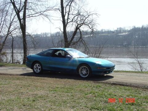 1993 Ford Probe - A 1993 Ford Probe GT sits parked by a river on a clear day, with trees and hills in the background.
