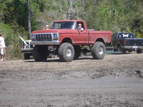 1989 Ford Bronco - A lifted red pickup truck with oversized tires navigates a muddy terrain, with another truck in the background.