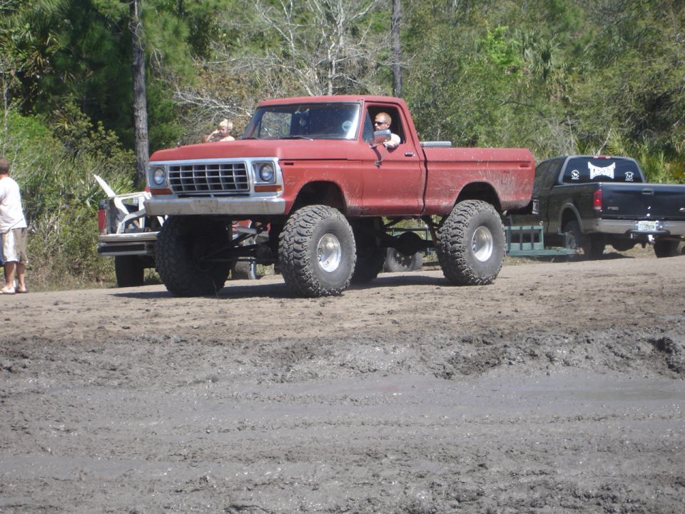 Lifted red pickup truck with large tires, driving in mud. Another black pickup truck is visible behind it