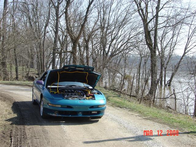Teal 1993 Ford Probe with hood open, parked by a flooded river