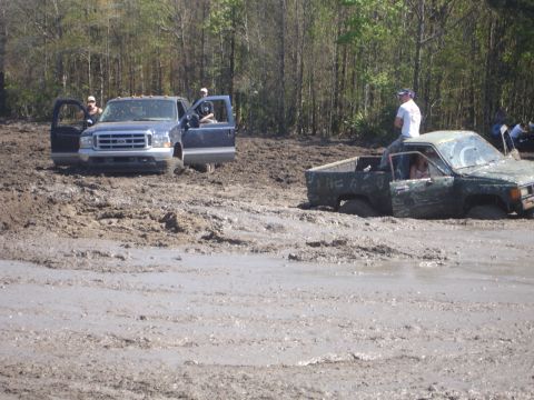 1989 Ford Bronco - Two trucks are stuck in a muddy area with trees in the background.