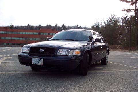 2001 Ford Crown Victoria - A dark blue 2001 Ford Crown Victoria police car is parked in an empty lot, facing the camera at a 3/4 angle.