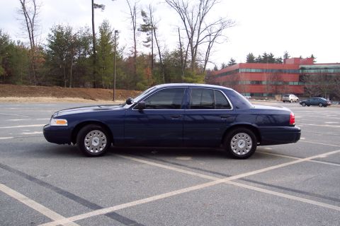 2001 Ford Crown Victoria - A dark blue 2001 Ford Crown Victoria is parked in an asphalt lot with trees and a building in the background.