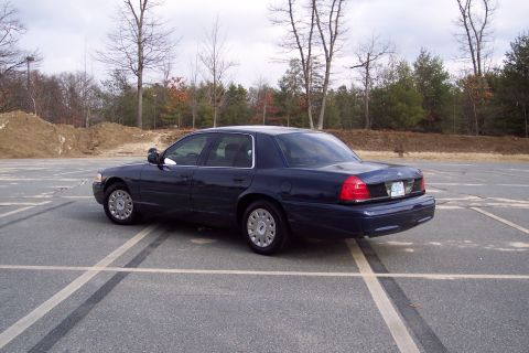 2001 Ford Crown Victoria - A dark blue 2001 Ford Crown Victoria sedan is parked in an empty lot under a cloudy sky.