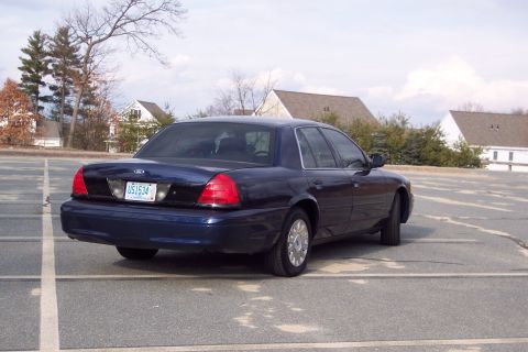 2001 Ford Crown Victoria - This dark blue sedan is parked in a lot with houses in the background.