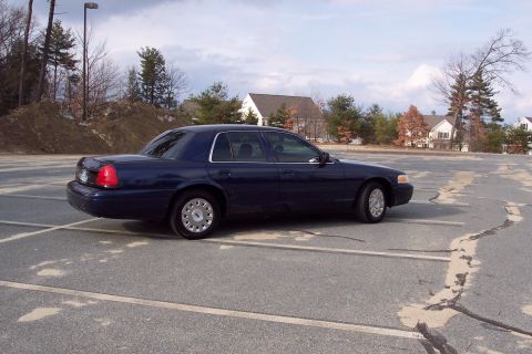 2001 Ford Crown Victoria - A dark blue sedan is parked in an empty lot under a cloudy sky.