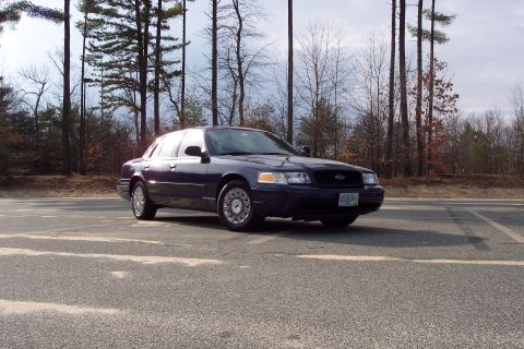 2001 Ford Crown Victoria - A dark blue 2001 Ford Crown Victoria is parked on an asphalt surface with a wooded area behind it.
