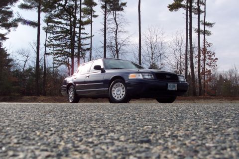 2001 Ford Crown Victoria - A dark blue Ford Crown Victoria is parked on the side of a road, with a backdrop of trees under a cloudy sky.