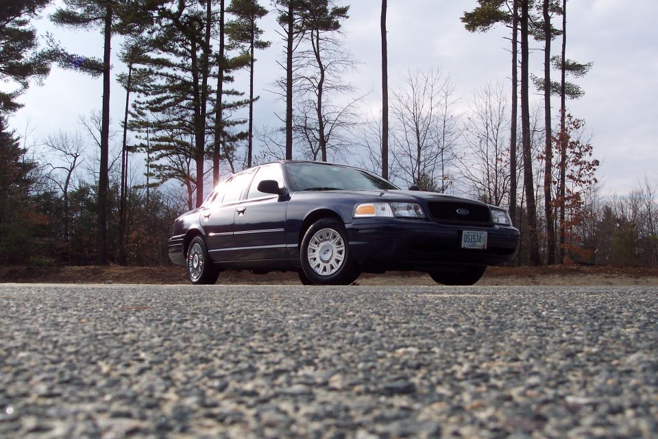 Dark blue Ford Crown Victoria parked on a road with trees in the background. License plate reads 051574