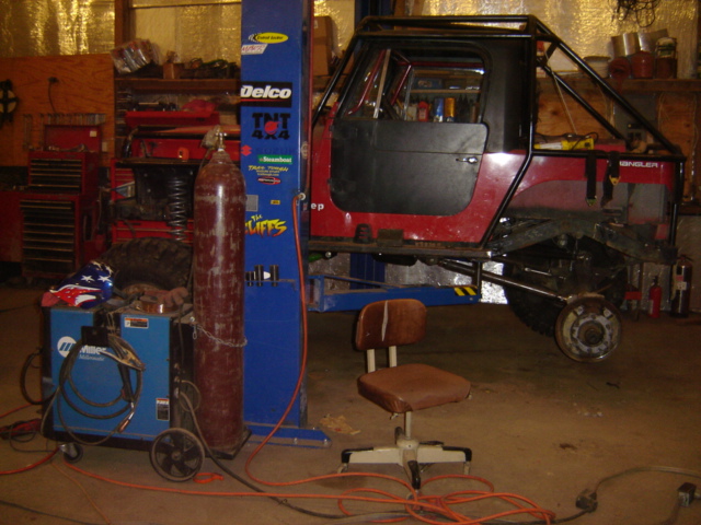 Red Jeep Wrangler on a lift in a garage with a welding machine and gas tank nearby