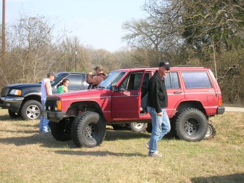 1996 Ford Bronco - A red Jeep Cherokee with modified off-road tires is parked on grassy terrain, with a black pickup truck visible behind it.