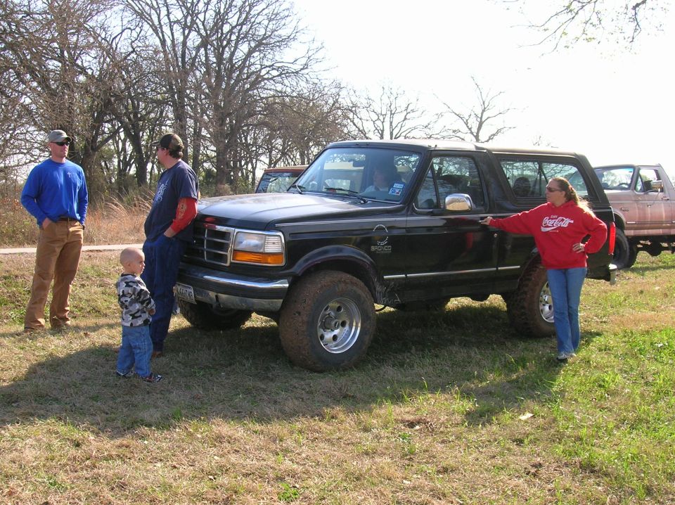 Black 1996 Ford Bronco parked in a grassy field with three people nearby; another truck is visible behind it