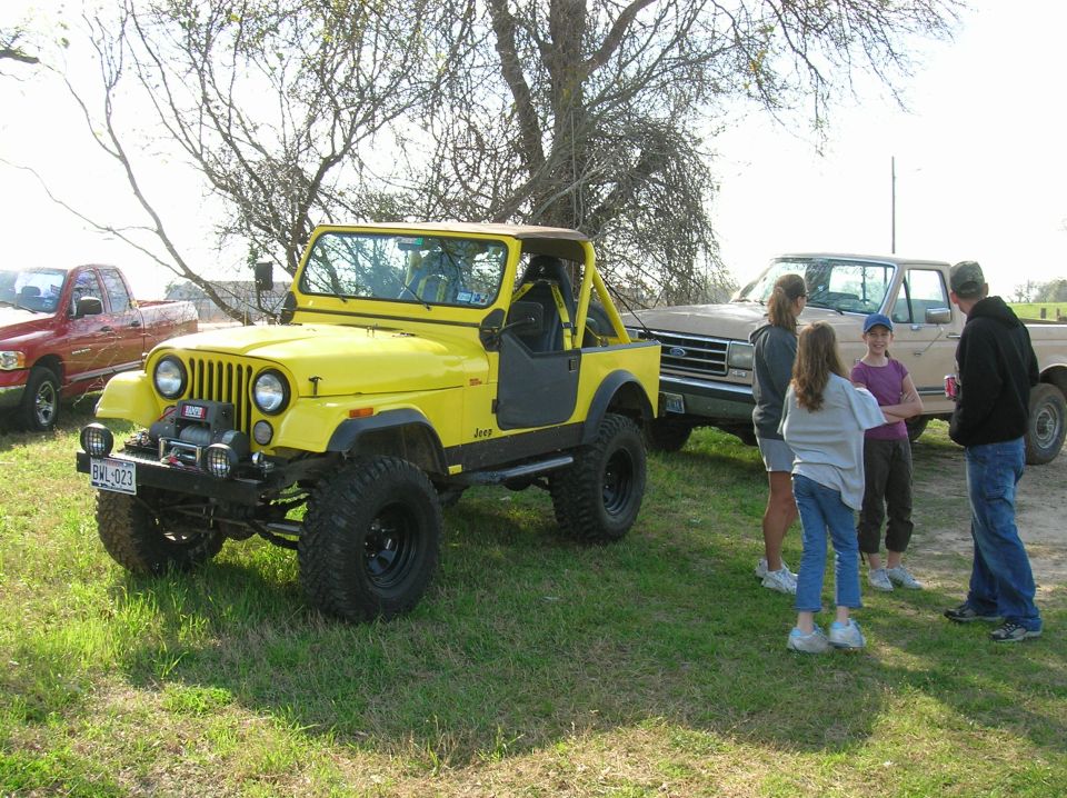 Yellow Jeep CJ with winch, parked next to a tan Ford pickup truck. Red pickup in background. People standing around