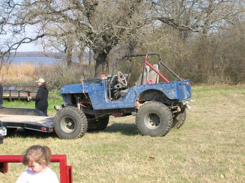1996 Ford Bronco - A modified blue jeep sits in a grassy field near a lake, with a trailer and a child visible in the foreground.