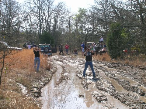 1996 Ford Bronco - Several off-road vehicles are stuck in deep mud on a wooded trail, with spectators observing.
