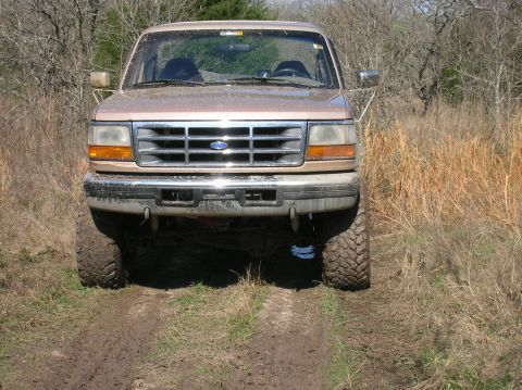 1996 Ford Bronco - A tan Ford Bronco with off-road tires drives through mud and dry grass.
