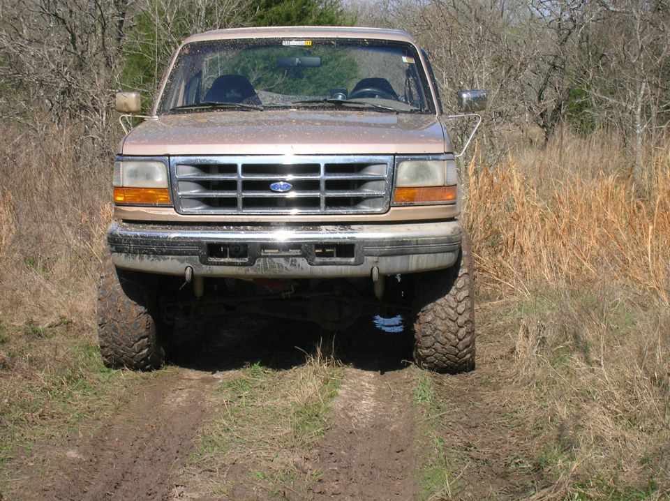 Tan Ford Bronco on muddy track, front view, off-road tires, trees and dry grass background