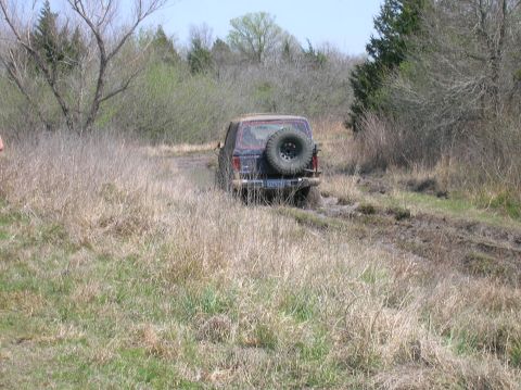 1996 Ford Bronco - An off-road vehicle navigates a muddy trail surrounded by dry grass and trees.