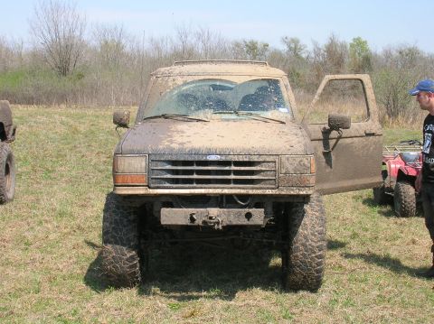 1996 Ford Bronco - A heavily mud-covered 1996 Ford Bronco sits in a field with its driver's door open, while another vehicle and an ATV are visible in the bac…