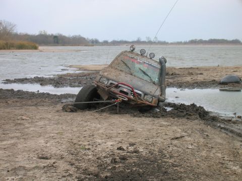 1996 Ford Bronco - A heavily mud-covered off-road vehicle is stuck on a muddy bank near a body of water, with a winch line attached.