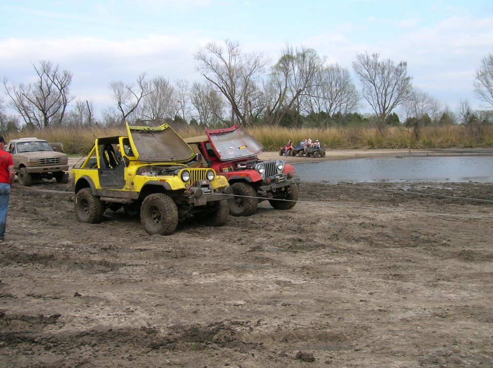 Muddy off-road vehicles with hoods open, one yellow Jeep and one red Jeep, near a body of water
