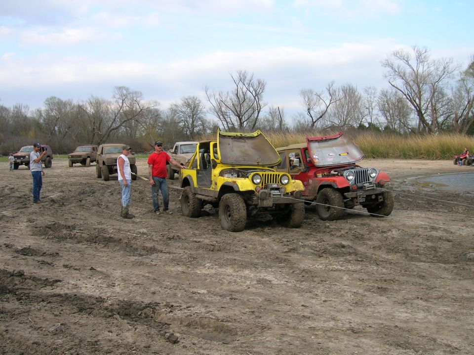 Yellow Jeep with hood open, being towed by red Jeep in muddy field