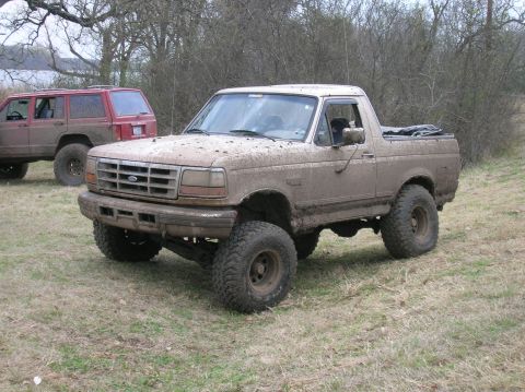 1996 Ford Bronco - A pair of off-road vehicles sit parked in a grassy field, covered in mud.