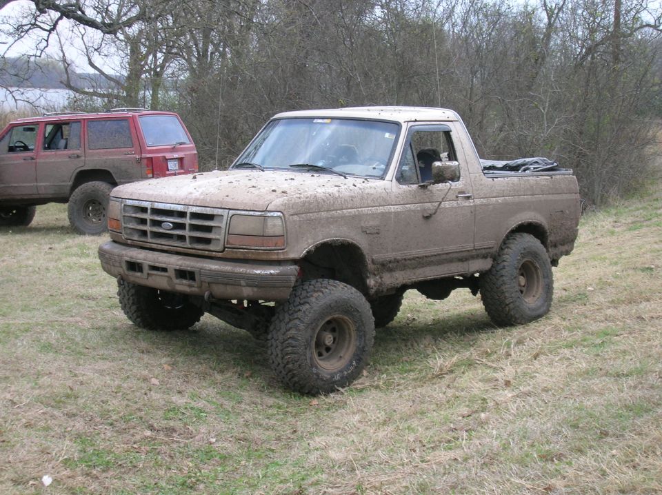 Muddy pickup truck with lifted suspension and off-road tires. Another red SUV is parked behind it