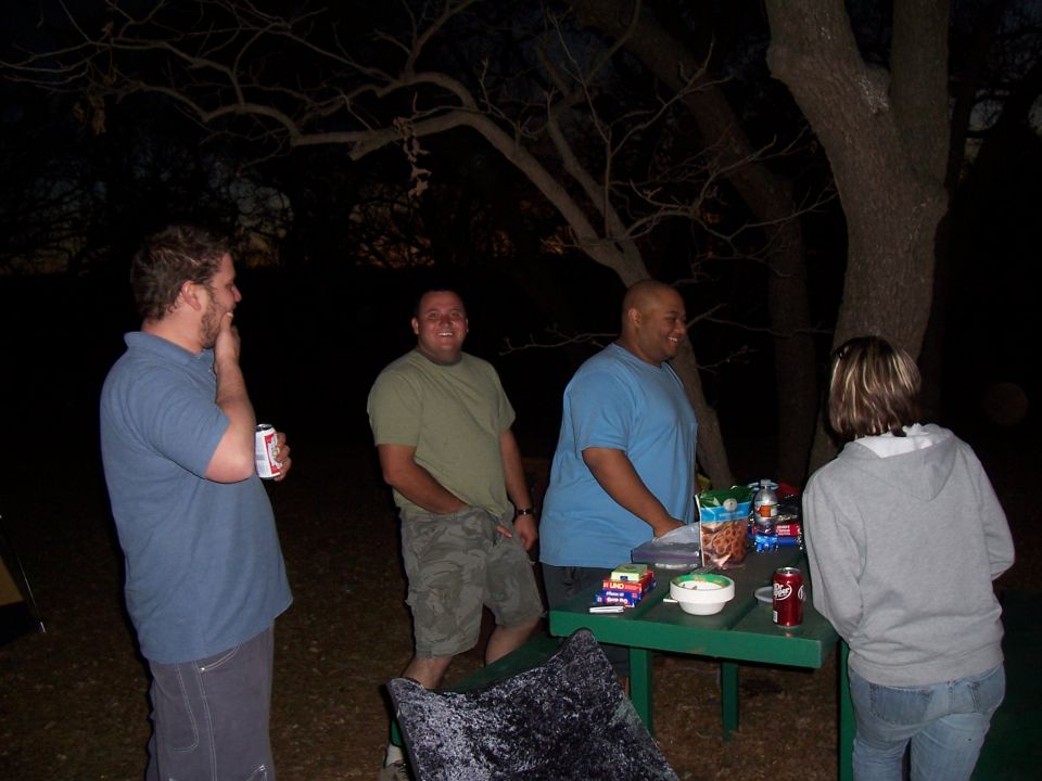 Four people stand around a picnic table outdoors at night, with snacks and drinks on the table