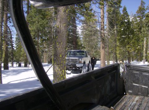 1986 Ford Bronco - A silver pickup truck with large tires is parked in a snowy forest with a person standing next to it.