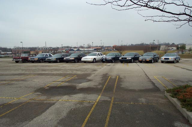 Row of full-size sedans parked side by side in a gravel lot under gray sky