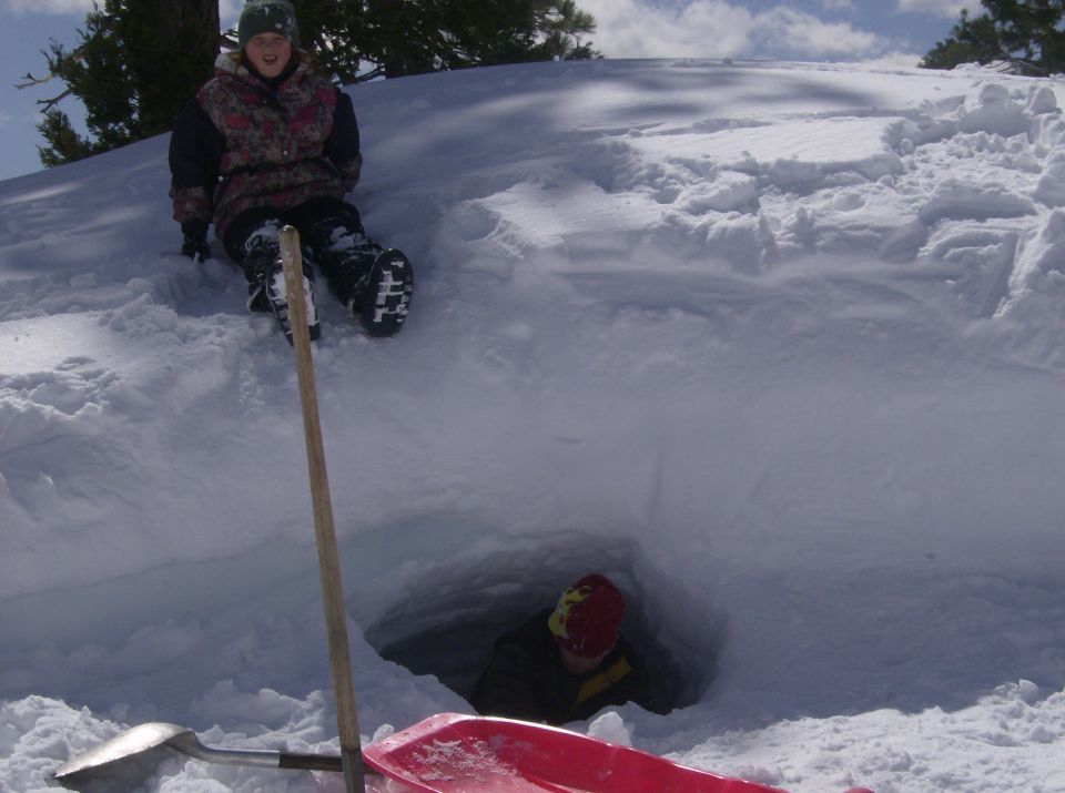 Kid sitting on top of snow fort, another kid inside. Shovel and sled nearby
