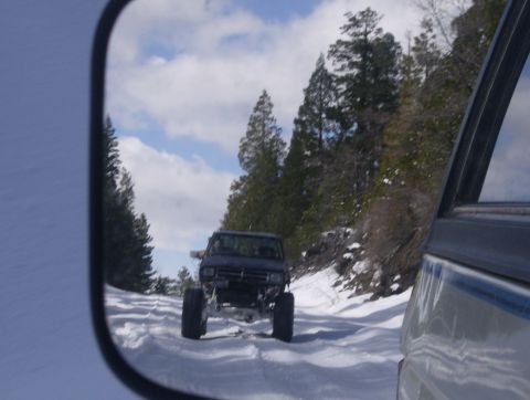 1986 Ford Bronco - A dark SUV with a lifted suspension navigates a snow-covered road, viewed from the side mirror of another vehicle.