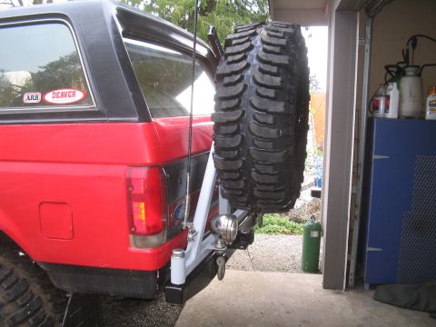 1989 Ford Bronco - The rear of a red Ford Bronco is shown with a spare tire mounted on a swing-away carrier. Two stickers are visible on the rear window.