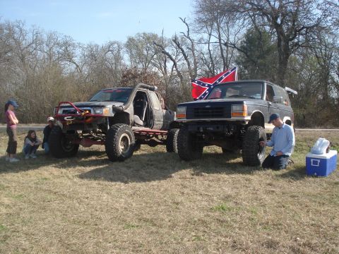 1989 Ford Bronco II - Two modified off-road vehicles are parked on a grassy field under a clear sky.