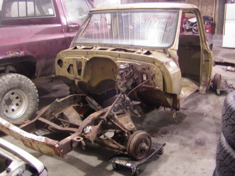 1991 Ford Bronco - A heavily rusted truck chassis and cab, stripped of its front end, sits in a garage next to another vehicle.