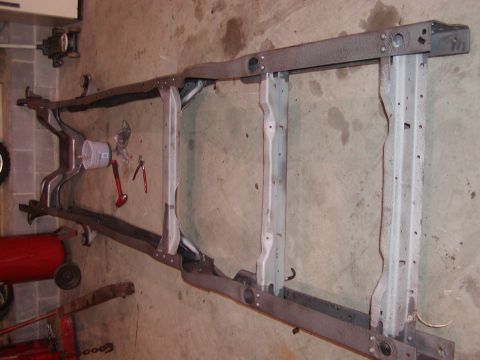 1991 Ford Bronco - A stripped ladder-type truck frame stood on edge against a block wall, showing the bare rails, several crossmembers, body mount holes and t…