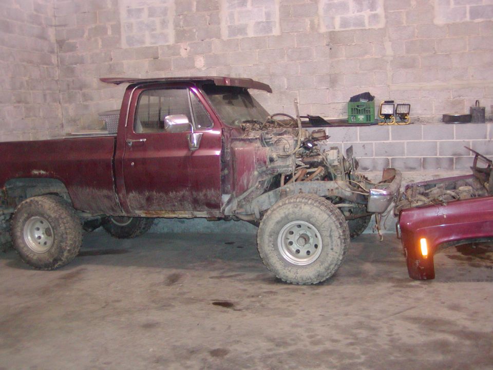 Maroon pickup truck frame with engine removed, side view. Another vehicle's front fender is visible