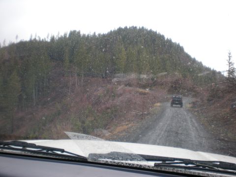 1993 Ford Bronco - Two SUVs travel on a wet gravel road surrounded by trees on a cloudy day.