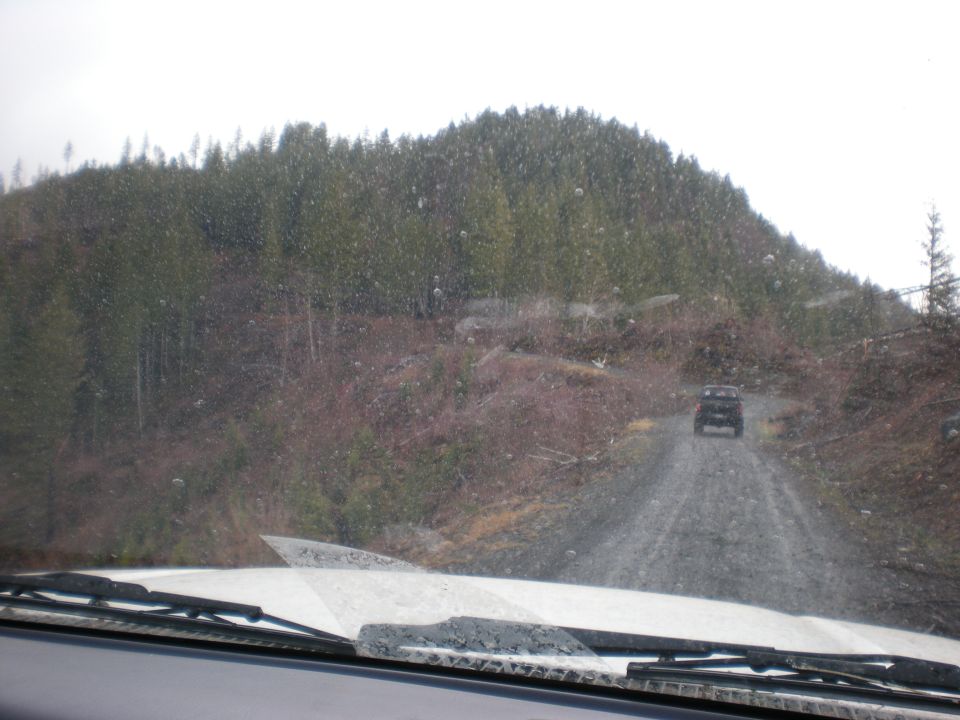 View from inside a vehicle looking at another SUV driving away on a wet gravel road through a forest