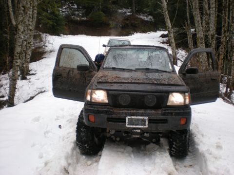 1993 Ford Bronco - A muddy black truck with its doors open sits in the snow, with another vehicle visible behind it on a wooded trail.