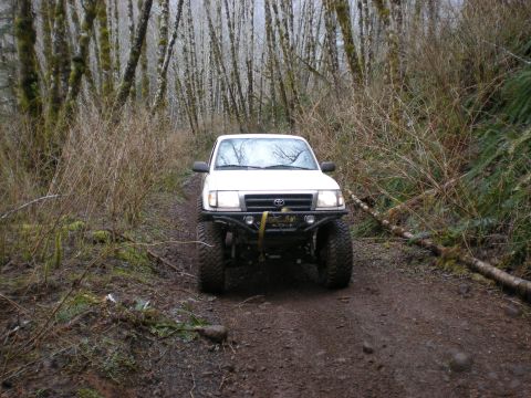 1993 Ford Bronco - A white Toyota Tacoma navigates a muddy trail surrounded by trees in a forest setting.