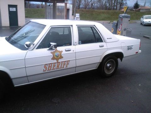 1991 Ford Crown Victoria - A white sheriff's car is parked outside, with a visible badge and 'SHERIFF' text on its side.