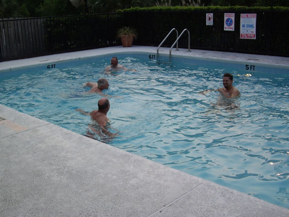 Four men swim in a backyard pool. Pool rules signs are visible on the fence