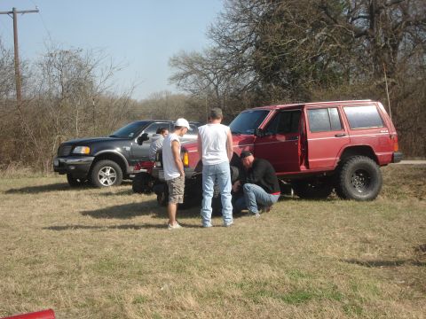 1989 Ford Bronco II - Several people gather around a red SUV, possibly working on a tire, with another vehicle in the background.