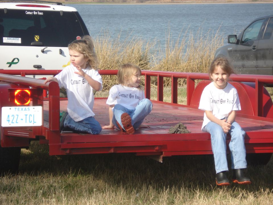 Three young girls sit on a red trailer. A white SUV with stickers is behind them, and a dark SUV is to the right. Water and dry grass are in the background