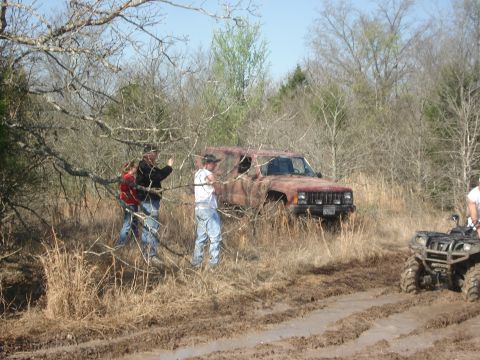 1989 Ford Bronco II - A group of people are gathered around a rusty Jeep stuck in a muddy, wooded area, with an ATV in the foreground.